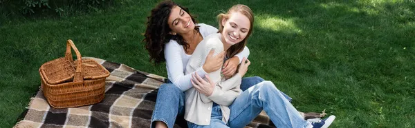 Fröhliche lesbische Frauen umarmen sich auf karierter Decke neben Korb, Banner — Stockfoto