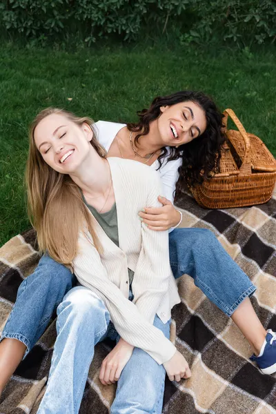 Visão de alto ângulo de mulheres lésbicas alegres abraçando e rindo no cobertor xadrez perto da cesta — Fotografia de Stock