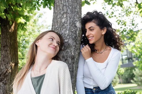 Happy lesbian women looking at each other near tree trunk outside — Stock Photo