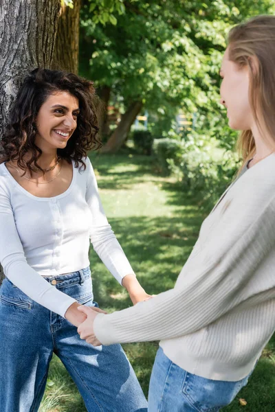 Pleased lesbian couple holding hands near tree trunk outside — Stock Photo