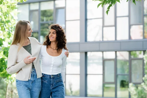 Smiling young woman hugging curly girlfriend while walking outside — Stock Photo