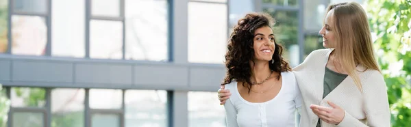 Lächelnde junge Frau umarmt lockige Freundin, während sie nach draußen geht, Banner — Stockfoto