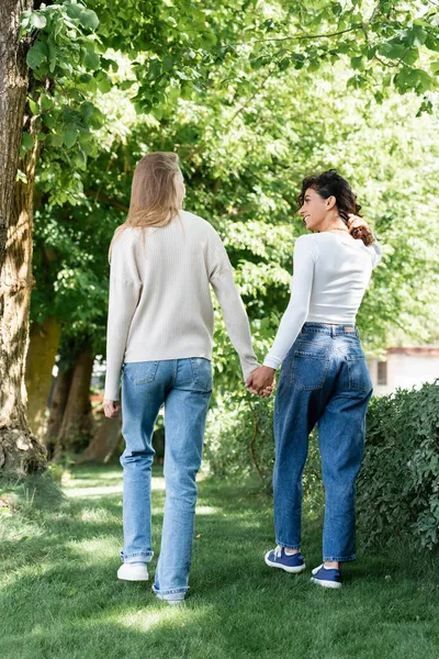 Full length of pleased lesbian couple in jeans holding hands and walking outside — Stock Photo