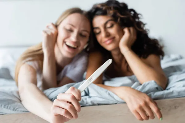Blurred and cheerful woman holding pregnancy test near curly girlfriend in bedroom — Stock Photo