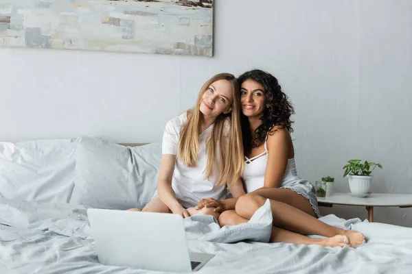 Pareja lesbiana positiva abrazándose mientras mira la cámara cerca de la computadora portátil en la cama - foto de stock