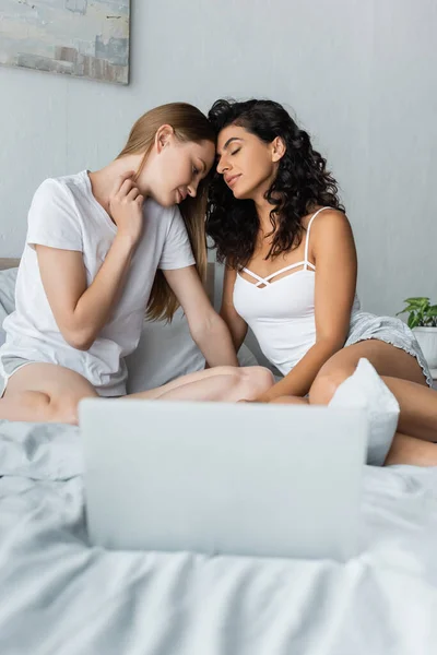 Loving same sex couple hugging near blurred laptop on bed — Stock Photo