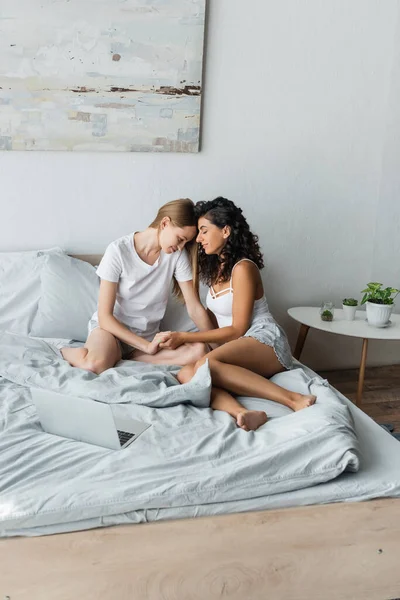 Amante del mismo sexo pareja cogida de la mano cerca de portátil en la cama - foto de stock