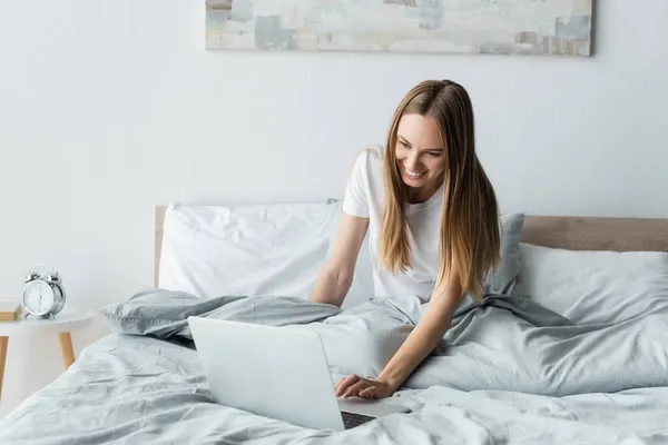 Happy young freelancer using laptop while sitting in bed — Stock Photo