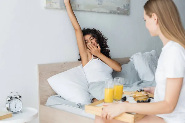 Junge Frau hält Tablett mit leckerem Frühstück in der Nähe lockige Freundin Stretching im Bett — Stockfoto