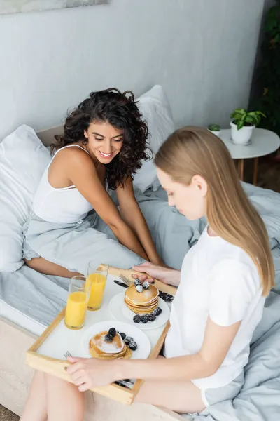 Prazer jovem segurando bandeja com saboroso café da manhã perto da namorada encaracolado na cama — Fotografia de Stock