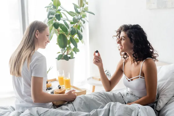 Mujer rizada sosteniendo mora y mirando a la novia feliz con bandeja - foto de stock