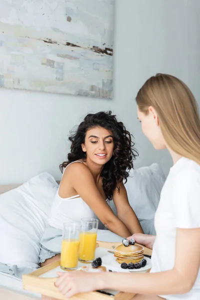 Jovem segurando bandeja com delicioso café da manhã perto de namorada encaracolado na cama — Fotografia de Stock