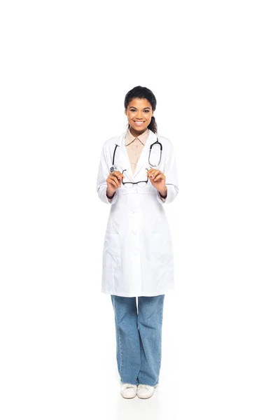 Full length of smiling african american doctor holding eyeglasses on white background — Stock Photo