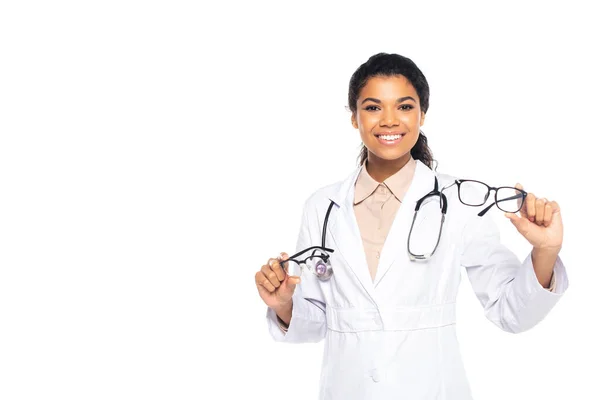 Positive african american ophthalmologist holding eyeglasses and smiling at camera isolated on white — Stock Photo