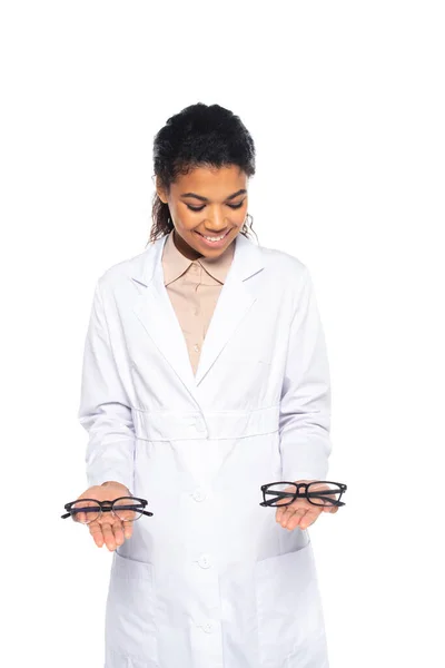 Happy african american oculist in white coat holding eyeglasses isolated on white — Stock Photo