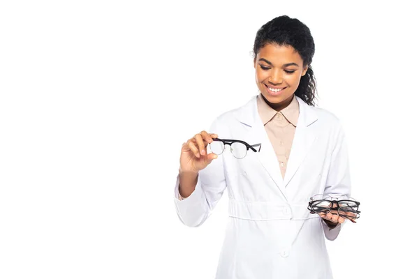 Ziemlich afrikanischer amerikanischer Augenarzt in weißem Mantel, der auf eine Brille blickt und isoliert auf weißem Grund lächelt — Stockfoto