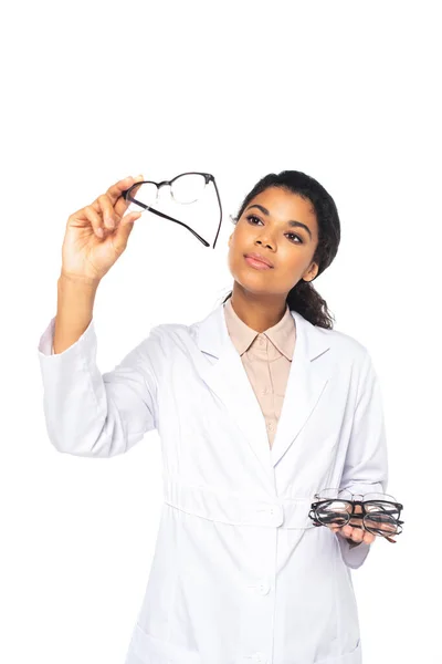 Jovem médico afro-americano segurando e olhando para óculos isolados em branco — Fotografia de Stock