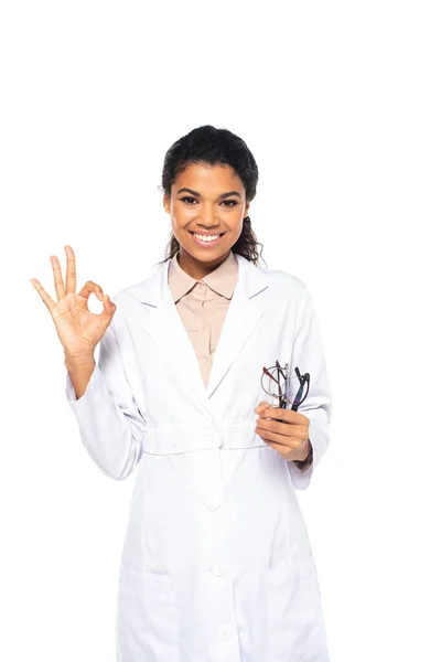 Cheerful african american oculist holding eyeglasses and showing ok gesture isolated on white — Stock Photo