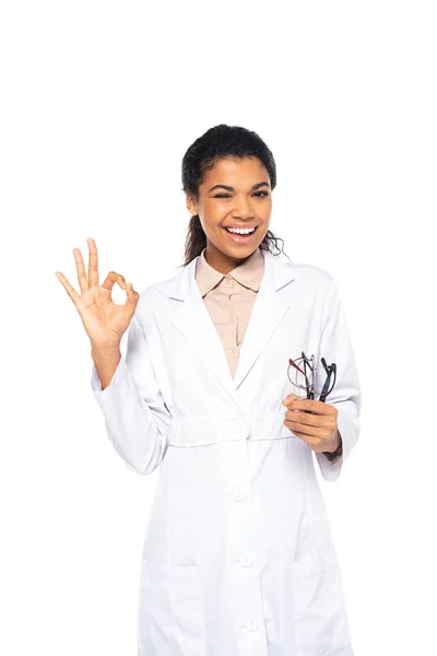 Happy african american oculist holding eyeglasses and showing ok gesture isolated on white — Stock Photo