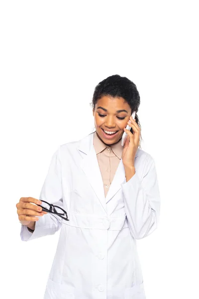 Excited african american oculist talking on smartphone and holding eyeglasses isolated on white — Stock Photo