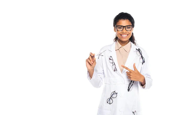 Positive african american oculist pointing at eyeglasses and looking at camera isolated on white — Stock Photo