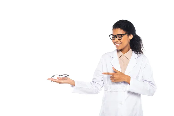 Smiling african american doctor pointing at eyeglasses on hand isolated on white — Stock Photo
