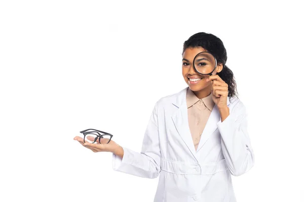 Smiling african american oculist holding eyeglasses and magnifying glass isolated on white — Stock Photo