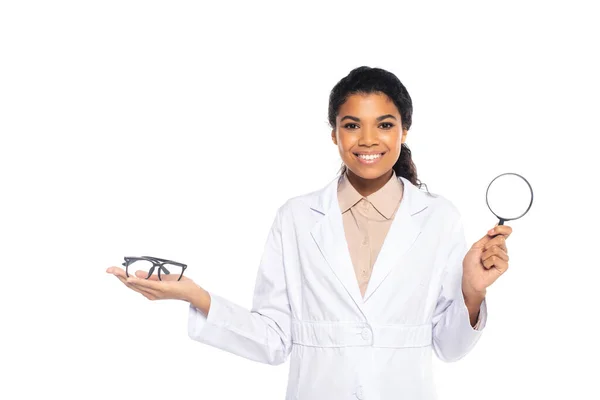 Cheerful african american doctor holding eyeglasses and magnifying glass isolated on white — Stock Photo