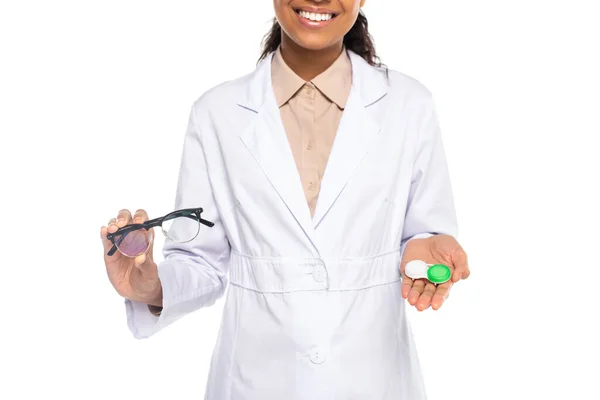 Vista recortada de um médico afro-americano sorridente segurando lentes de contato e óculos isolados em branco — Fotografia de Stock