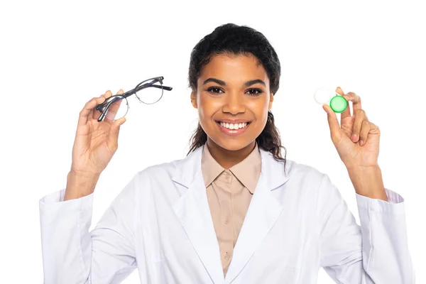 Pretty african american doctor holding optical eyeglasses and contact lenses in container isolated on white — Stock Photo