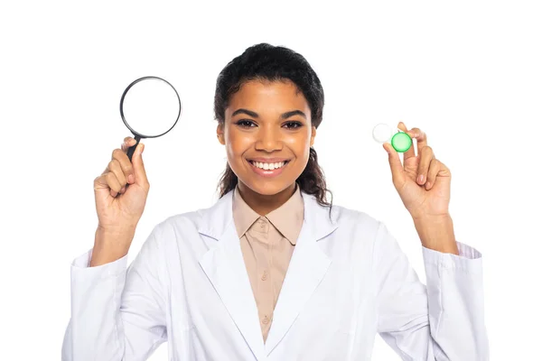 Smiling african american doctor holding magnifying glass and contact lenses isolated on white — Stock Photo