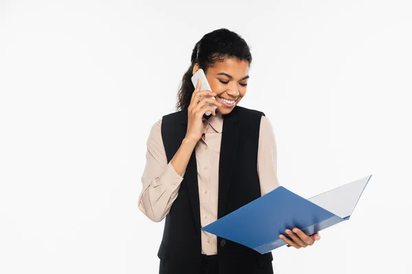Alegre mujer de negocios afroamericana hablando por teléfono móvil y sosteniendo la carpeta de papel aislada en blanco - foto de stock