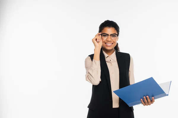 Cheerful african american businesswoman in vest holding eyeglasses and paper folder isolated on white — Stock Photo