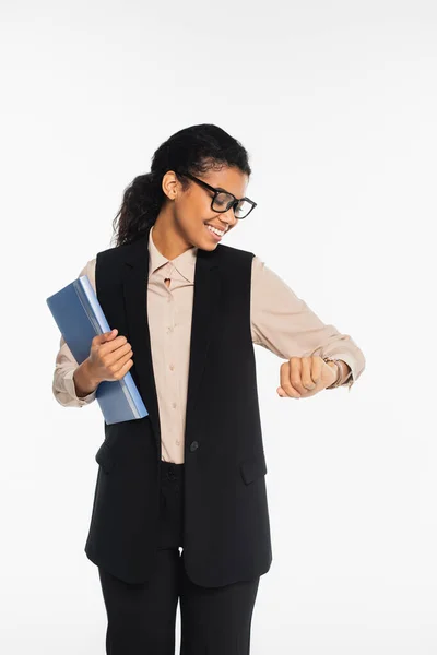 Empresaria afroamericana positiva sosteniendo carpeta de papel y mirando reloj de pulsera aislado en blanco - foto de stock