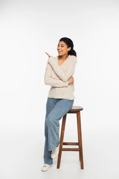 Souriante femme afro-américaine pointant du doigt près de la chaise sur fond blanc — Photo de stock