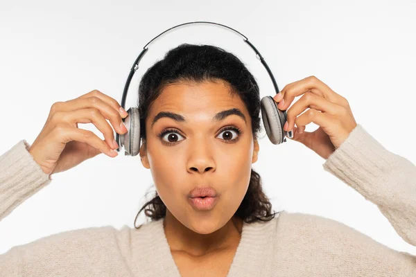 Mujer afroamericana asombrada sosteniendo auriculares y mirando a la cámara aislada en blanco - foto de stock