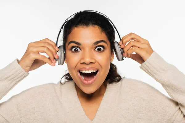 Mulher afro-americana excitada em suéter segurando fones de ouvido perto de orelhas isoladas em branco — Fotografia de Stock