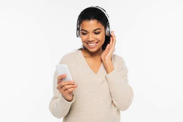Alegre mujer afroamericana en suéter usando auriculares y teléfonos celulares aislados en blanco — Stock Photo