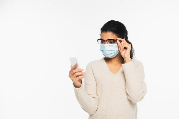 Jovem afro-americana em máscara médica e óculos usando smartphone isolado em branco — Fotografia de Stock