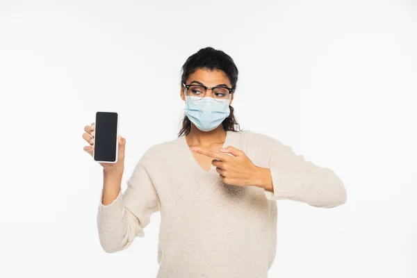 African american woman in eyeglasses and medical mask pointing at smartphone isolated on white — Stock Photo