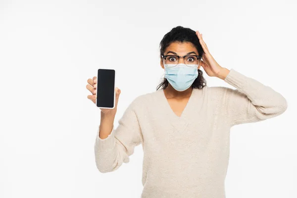 Mujer afroamericana asombrada en gafas y máscara médica sosteniendo teléfono inteligente aislado en blanco - foto de stock