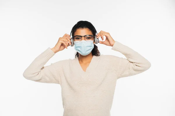 Mulher afro-americana em óculos e máscara médica usando fones de ouvido isolados em branco — Fotografia de Stock