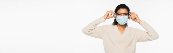 Femme afro-américaine en lunettes et masque médical à l'aide d'écouteurs isolés sur blanc, bannière — Photo de stock