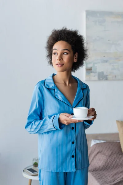 Ricci donna afroamericana in possesso di una tazza di caffè e piattino in camera da letto — Foto stock