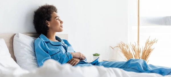 Glückliche afrikanisch-amerikanische Frau im Schlafanzug auf dem Bett liegend, Banner — Stockfoto