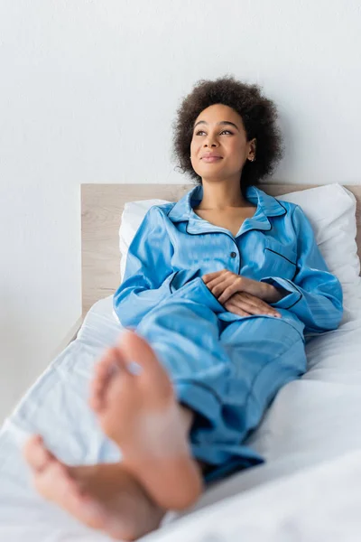 Felice donna afroamericana in pigiama poggiata sul letto — Foto stock