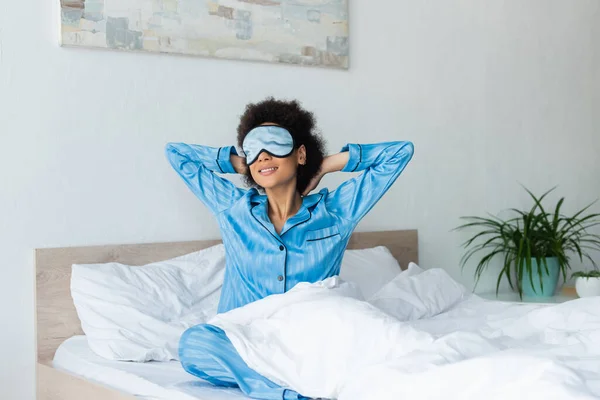 Весела афроамериканська жінка в піжамі і спляча маска сидячи на ліжку. — Stock Photo
