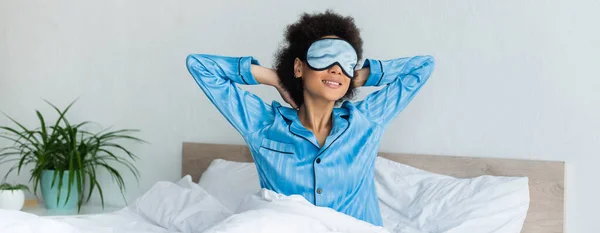 Alegre afroamericana mujer en pijama y dormir máscara sentado en la cama, bandera - foto de stock