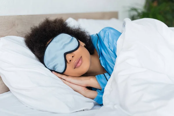 Femme afro-américaine bouclée en pyjama bleu et masque pour les yeux en soie dormant au lit — Photo de stock