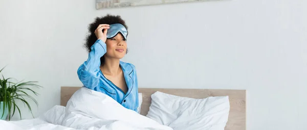 Somnolienta mujer afroamericana en pijama ajustando la máscara de dormir, pancarta - foto de stock